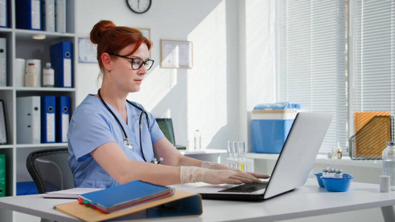 The doctor is sitting at her desk, working on her laptop