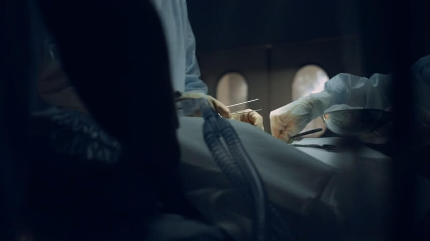 A dimly lit operating room where two surgeons in protective gowns and gloves perform a delicate procedure on a patient. The scene conveys the seriousness of surgical risks