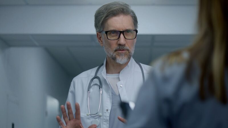 A senior male doctor with gray hair, glasses, and a stethoscope around his neck is speaking to a colleague in a hospital corridor