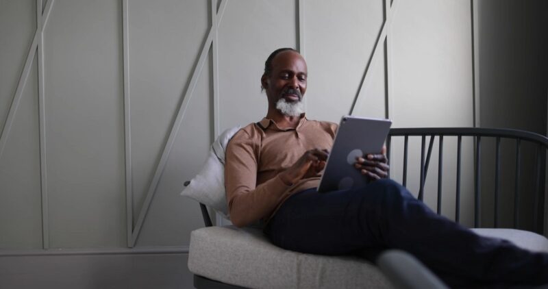 An older man with a salt-and-pepper beard reclines on a couch, using a tablet