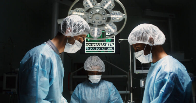 Three surgeons in an operating room, dressed in full surgical gear including masks and hairnets, working under bright overhead lights. 