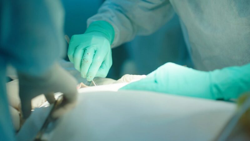 A surgeon wearing green gloves carefully sutures a patient's incision under bright surgical lights.