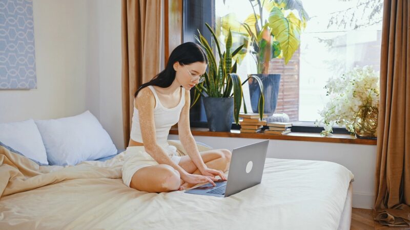 A woman with glasses and vitiligo sits cross-legged on a bed, typing on a laptop